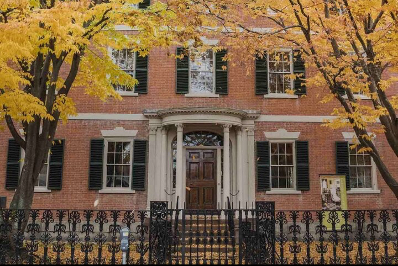A brick building with black shutters is framed by yellow-leaved trees in autumn. The entrance has a columned portico and a dark wooden door. A black iron fence runs along the front. Leaves are scattered on the ground.