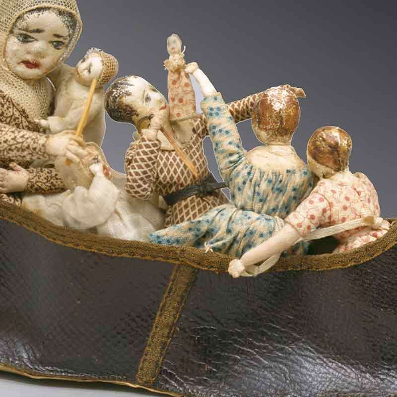 A group of five handmade dolls with painted faces and patterned clothing sits together in a brown, boat-shaped vessel. One doll is holding a small stick and another is raising a tiny doll in the air.