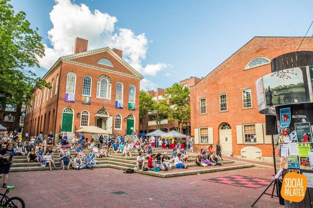 A crowd of people sits and stands on brick steps in a public square surrounded by historic red brick buildings on a sunny day. A sign in the foreground reads "Social Palates.