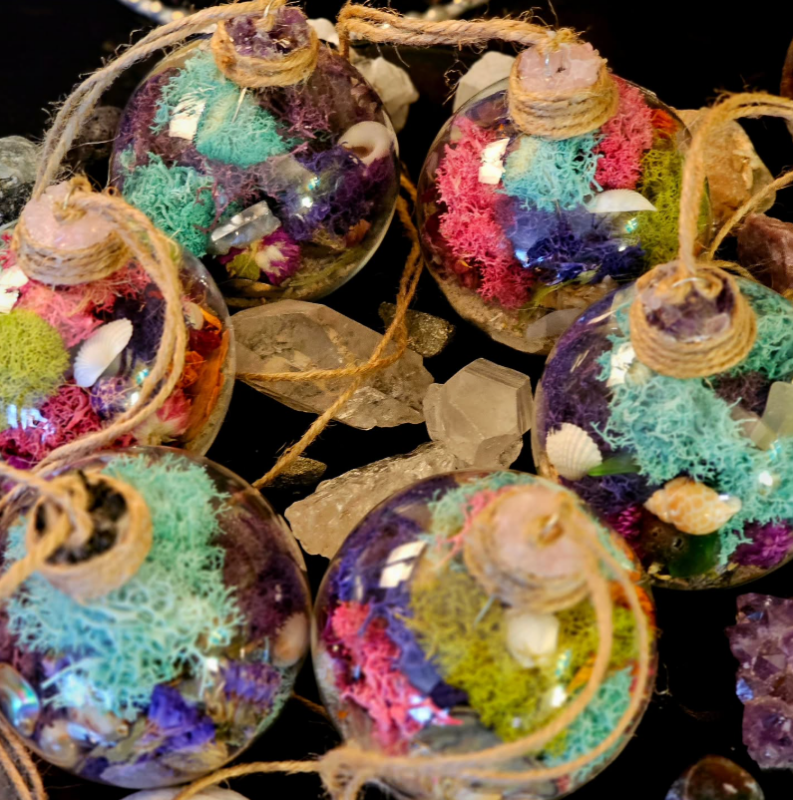 Six glass ornaments filled with colorful dried moss, small seashells, and decorative stones are arranged on a surface with clear crystals scattered among them. Each ornament has a jute string for hanging.