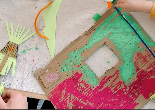 A child’s hands work on a cardboard craft painted with green and red colors, using a blue pipe cleaner. In this K-2 Crafts Class, green paper pieces and a pair of scissors are also visible on the table, sparking Eco Arts creativity.