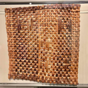 A large, rectangular woven textile made from various shades of brown material. The uneven weaving pattern and checkerboard appearance evoke the rustic charm of an April vacation in Essex County. Displayed against a light wall.