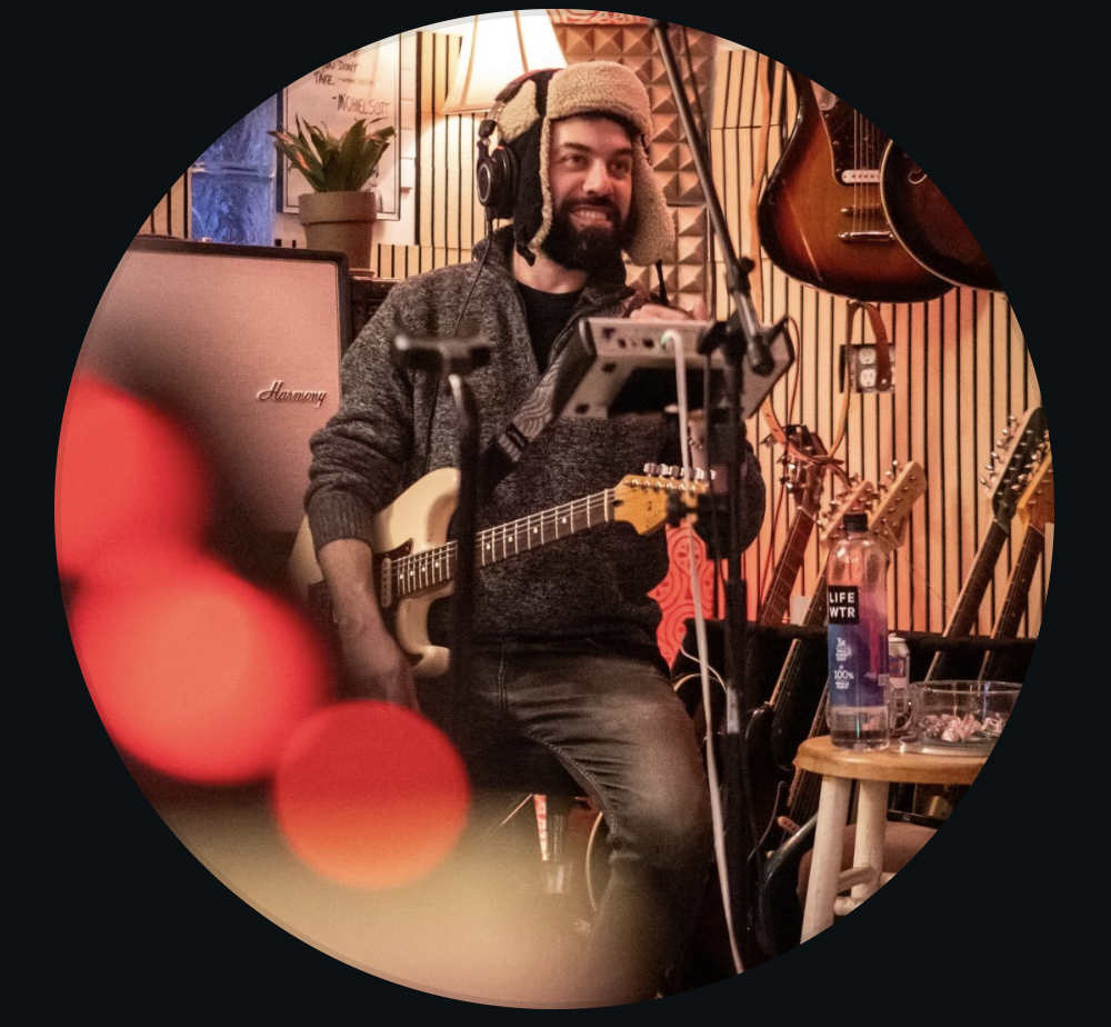 A man wearing a winter hat and headphones sits in a music studio holding an electric guitar, ready for live music. He is surrounded by guitars, an amplifier, a stand with a tablet, and a water bottle—true Reilly Somach style.
