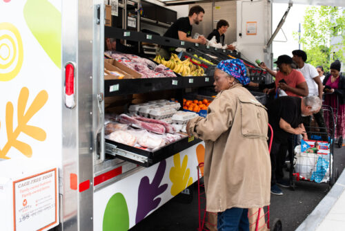 Several people shop for groceries at an outdoor market stand, selecting items such as fruits, vegetables, and packaged goods from shelves on a colorful truck, with employees assisting behind the counter.