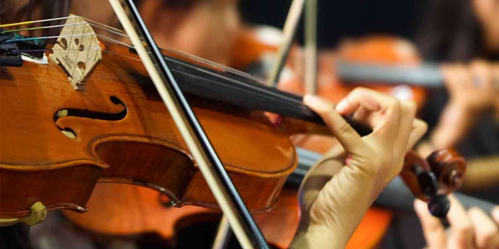 Close-up of a person playing a violin with a bow, focusing on the musician’s hand and the instrument’s strings. Other violinists are visible in the blurred background.