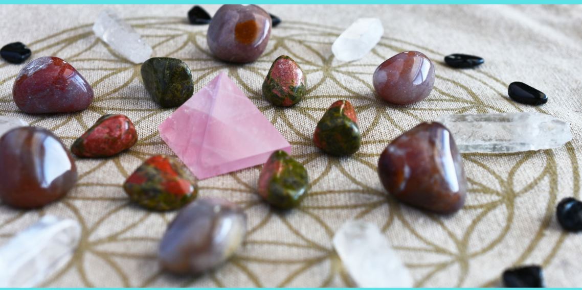 A group of assorted tumbled stones and crystals arranged in a circular pattern on a beige cloth with a geometric design, featuring a pink pyramid-shaped crystal in the center.