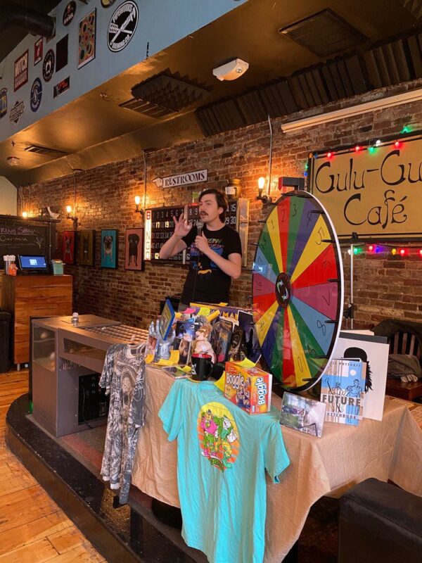 A person stands behind a table covered with prizes and a colorful spinning wheel in a café with brick walls, string lights, and signs. T-shirts, board games, and books are displayed on the table.
