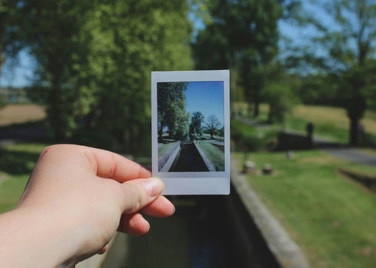 A hand holds a Polaroid photo showing a tree-lined canal—an example of photography capturing a moment in time. The background matches the Polaroid image, depicting the same canal, trees, and grassy area under a clear blue sky.