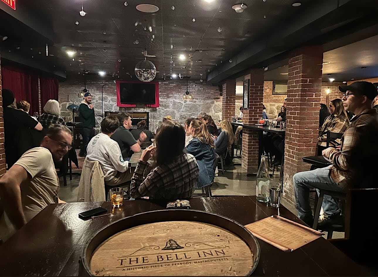 A large group of people sit and stand around tables in a dimly lit bar with brick columns. A person is speaking at a microphone in front of a red curtain. The foreground shows a round table labeled "The Bell Inn.