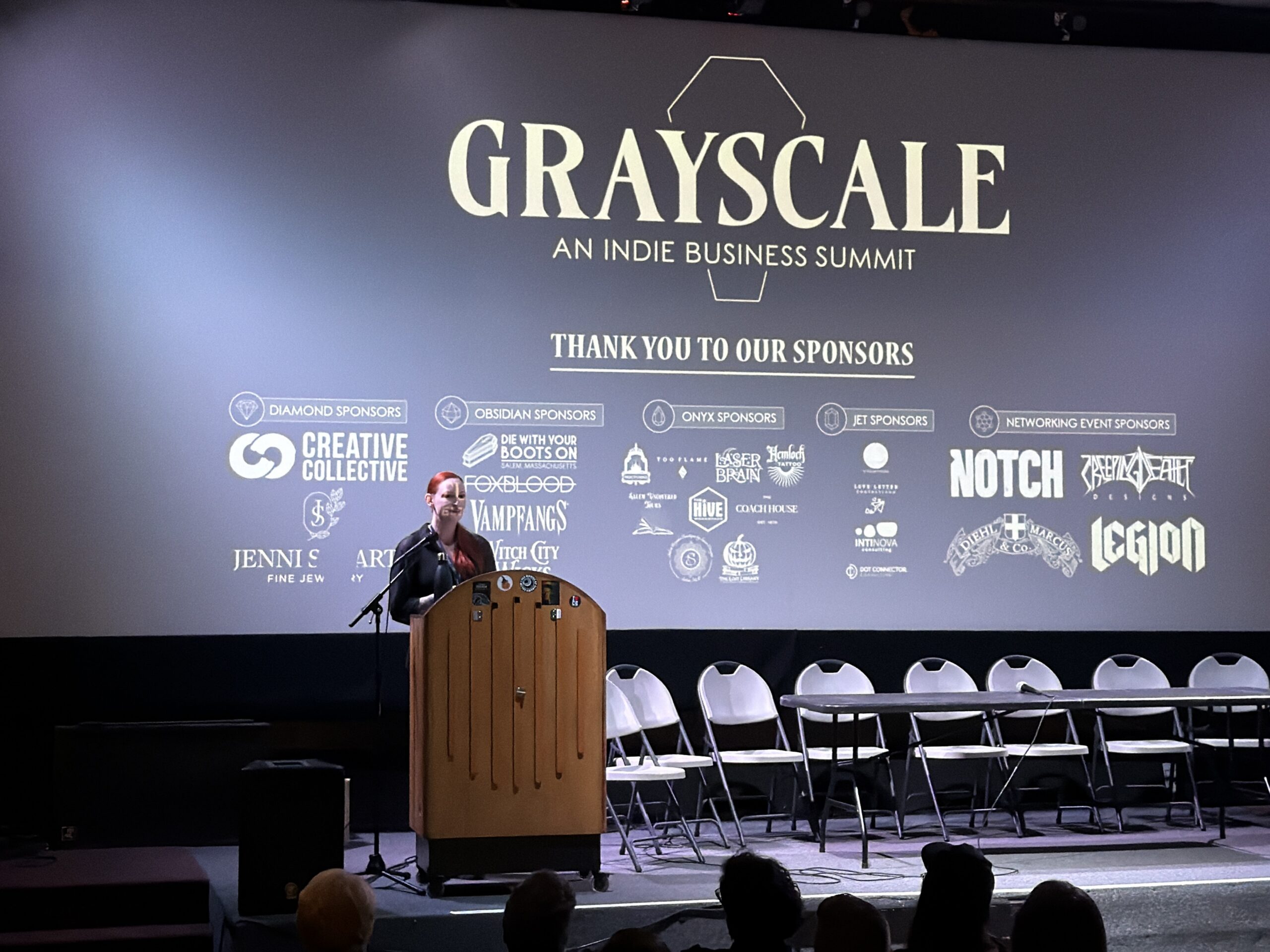 A person stands at a podium on stage in front of a large screen displaying the title "Grayscale: An Indie Business Summit," a list of sponsors, and six empty chairs—ready for a panel on economics and thriving In the Black.