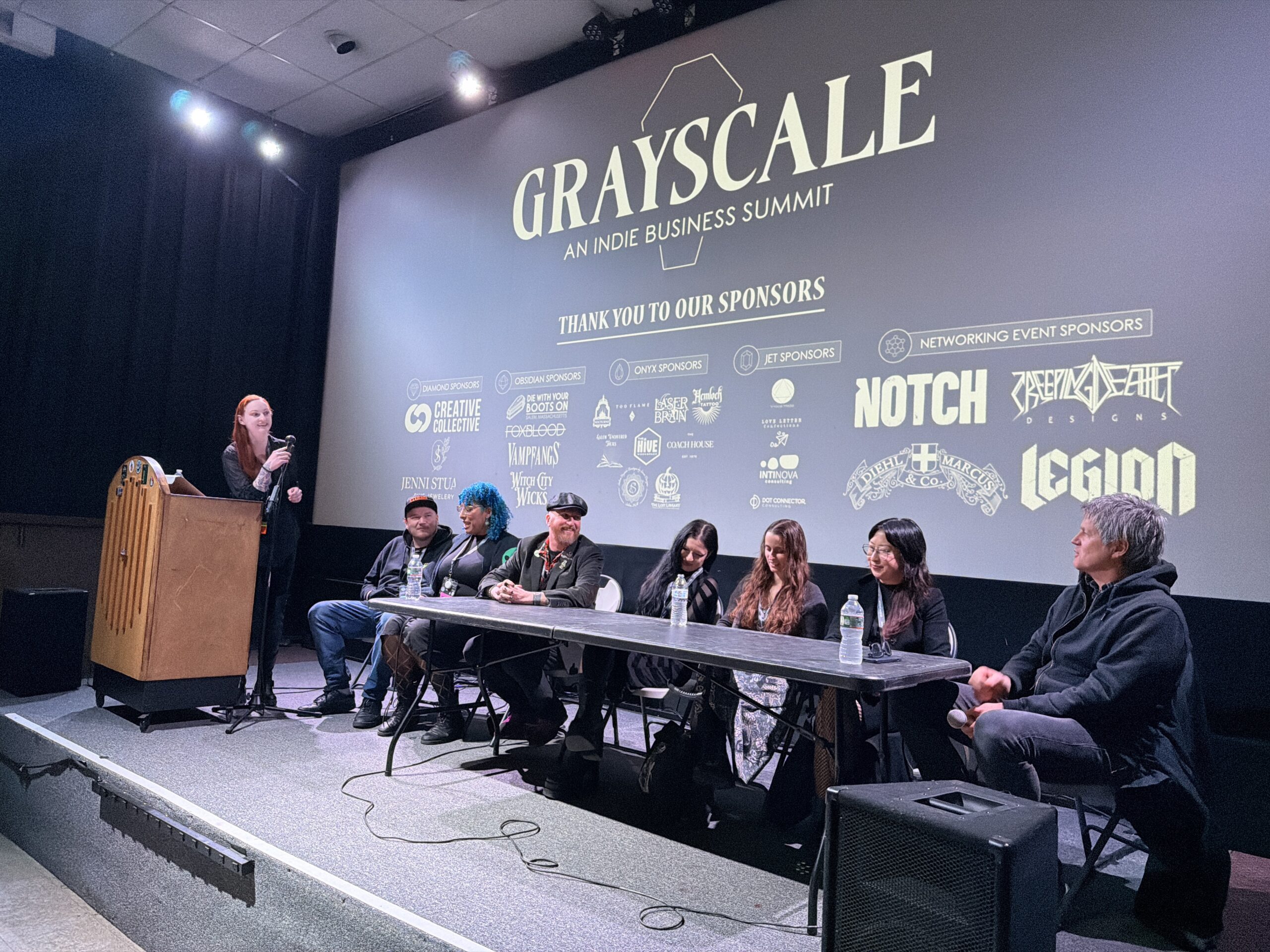 A group of seven people sits at a long table on stage during a panel at Grayscale: An Indie Business Summit. As discussions on economics unfold, a person stands at the podium, with sponsor logos and "In the Black" projected behind them.