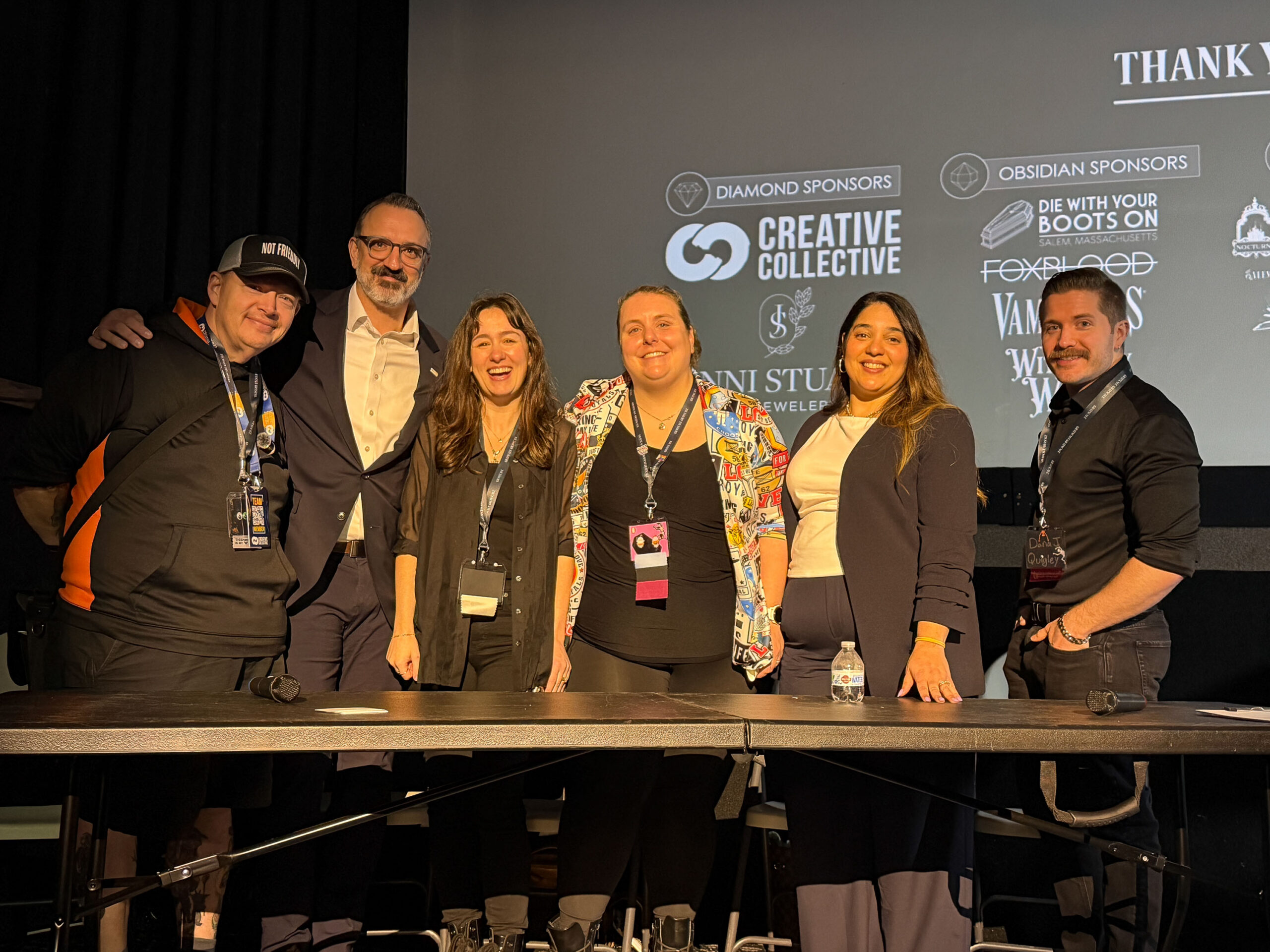 Six people stand behind a table on stage, posing for a group photo at an Economics event. Behind them, a projected screen displays sponsor logos and the words "Thank You." Water bottles and microphones are on the table in front.