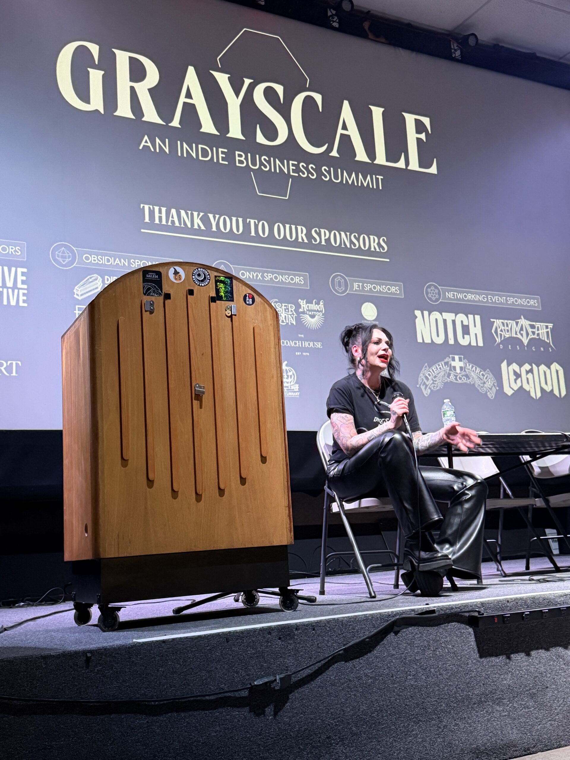 A person sits on stage at the Grayscale Indie Business Summit, speaking on economics into a microphone. Sponsor logos are displayed behind them, with a wooden podium and table also on stage—a scene where business meets the weird.