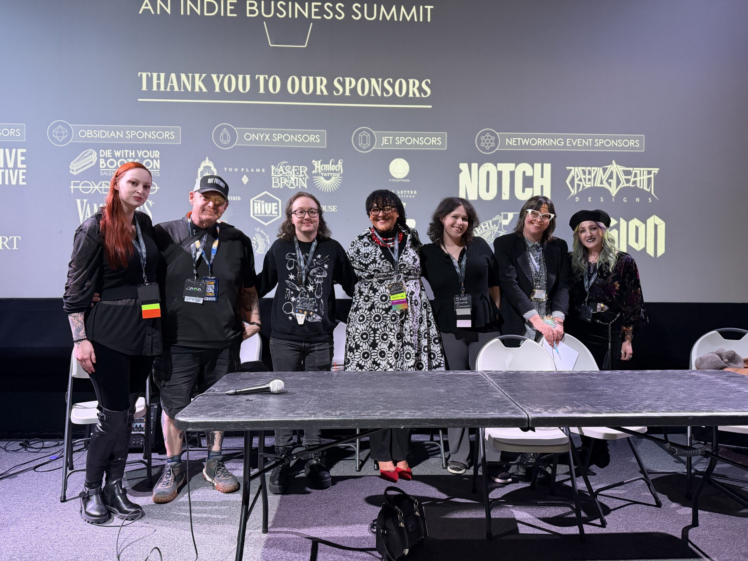 Seven people stand side by side on stage behind a table, posing for a group photo at the In the Black indie business summit. A sponsor thank you slide is projected on the screen behind them, highlighting their support of creative economics.
