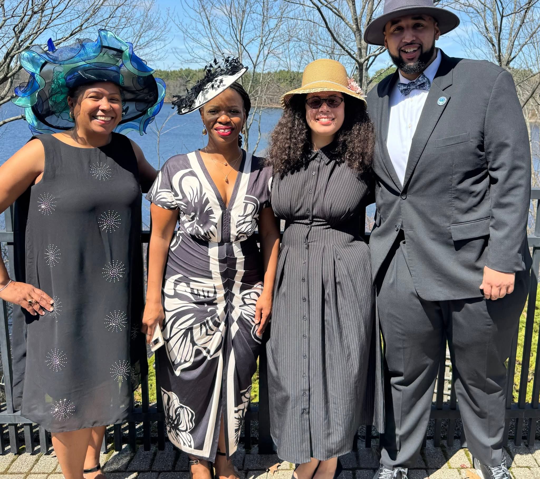 Four adults stand side by side outdoors at a North Shore Tea Event in sunny weather, wearing formal attire and decorative hats, with a body of water and leafless trees visible in the background.