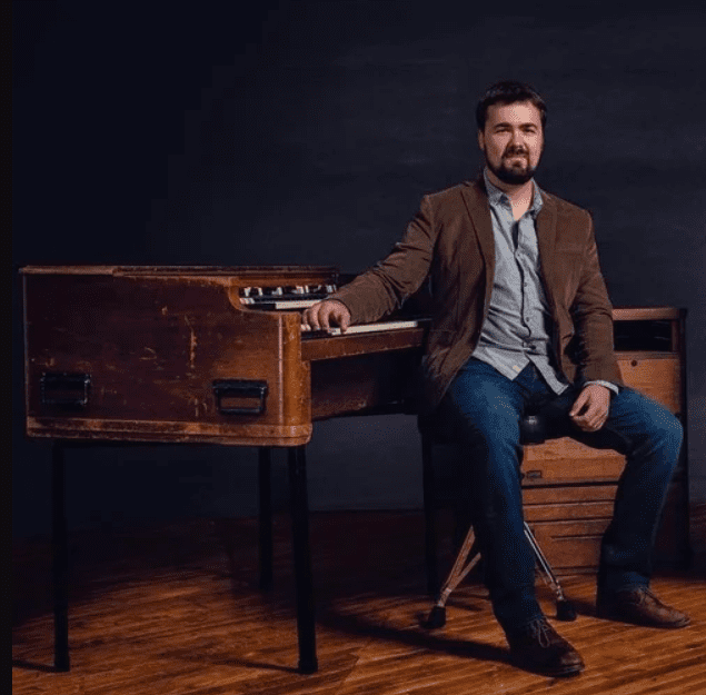 A man with dark hair and a beard sits on a stool next to a vintage wooden organ, resting one hand on the keyboard. He is wearing a brown jacket, blue jeans, and brown shoes, with a dark background behind him.
