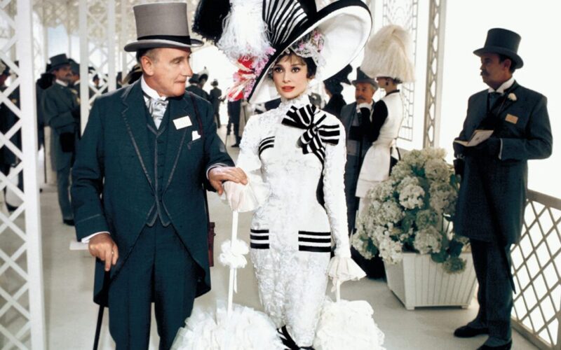 A woman in an elaborate white dress with black stripes and a large hat stands next to a man in a dark suit and top hat. Several other men in formal attire stand in the background at an ornate, white venue.