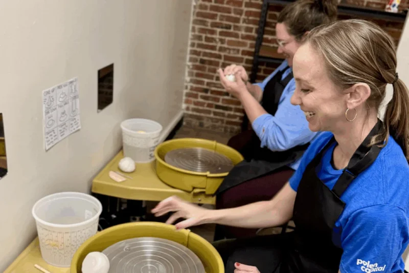 Two women participate in a pottery workshop, shaping clay balls at their wheels. One smiles as she prepares to begin, while the other works with clay in her hands. Both wear aprons in an inviting Adult Claymaking studio with brick and plain walls.