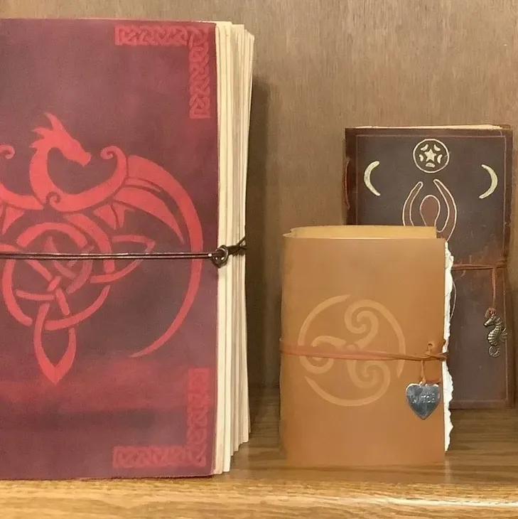 Three leather-bound journals with decorative covers, crafted at a bookbinding workshop, sit on a wooden shelf. The largest is maroon with a dragon design, the middle is tan with a swirl, and the smallest is brown with moon and star symbols.