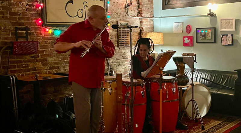 A man in a red shirt plays a flute while a woman sits behind red conga drums reading sheet music. They are performing World Music in a cozy, brick-walled café adorned with colorful lights and framed art.