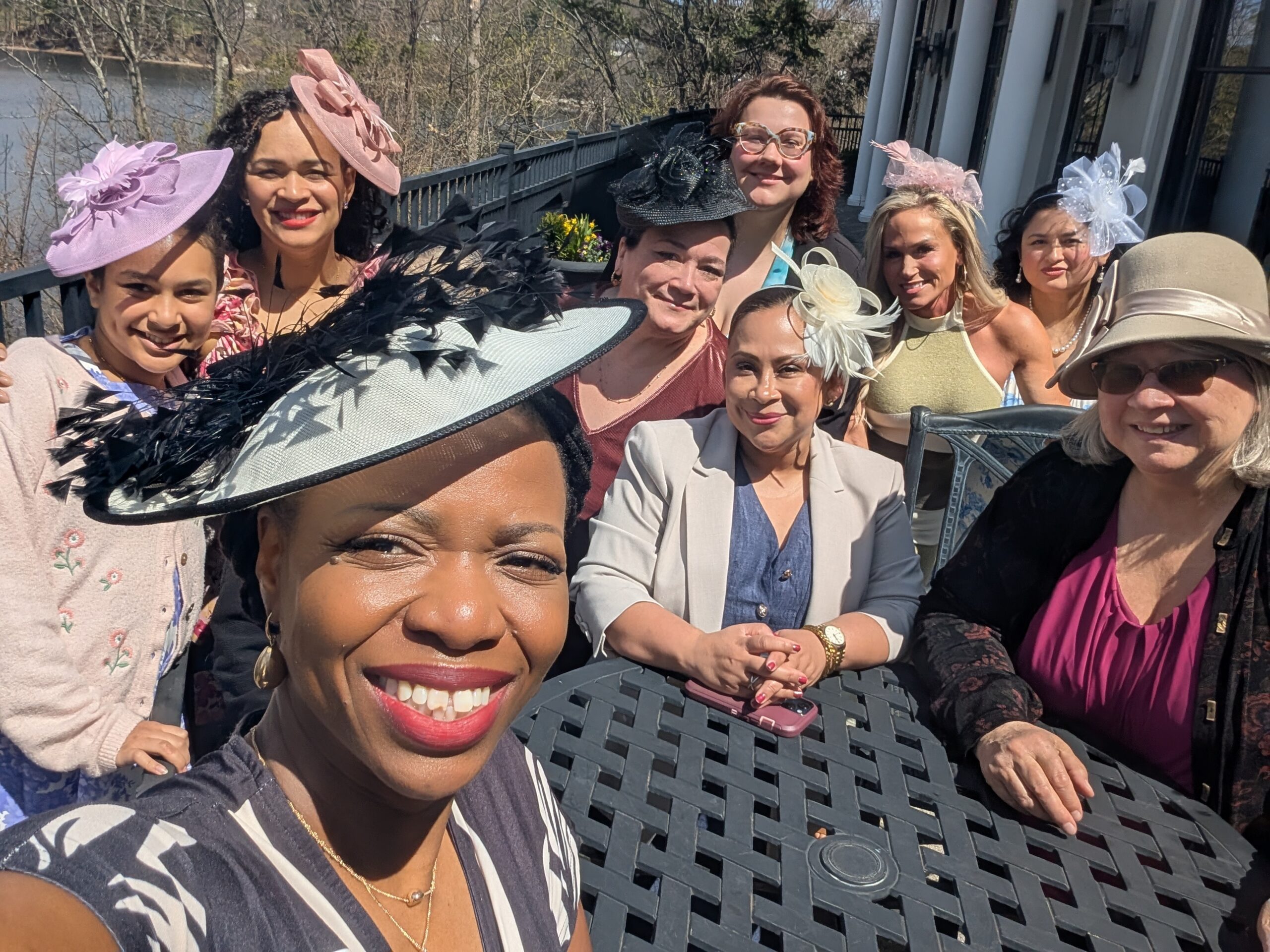 A group of nine women in hats and spring attire pose for a selfie at the North Shore Juneteenth Association Tea, seated and standing around a black metal table outdoors with trees and a river in the background.