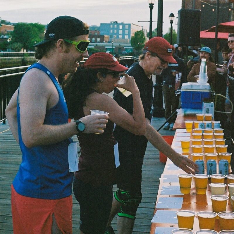 Three runners wearing race bibs and athletic gear stand at a table lined with cups of beer, drinking and preparing to pick up more, during a daytime outdoor event on a boardwalk.