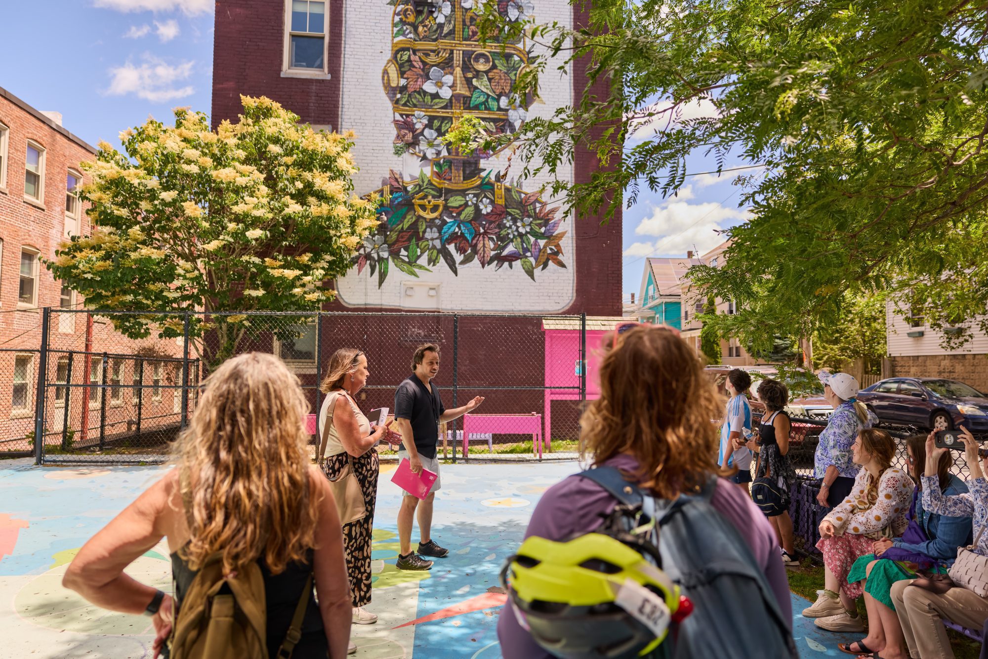 A group of people stands outdoors in front of a large, colorful mural painted on a brick building. One person appears to be speaking to the group. Trees and a chain-link fence are visible nearby.