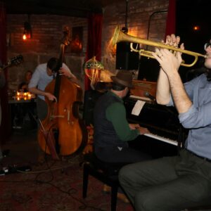A jazz band performs indoors for the Sevenings: About Time Edition. One musician plays trumpet in the foreground, another upright bass, a third piano, and a fourth is partially visible on guitar. The setting features dim lighting, brick walls, and red curtains.
