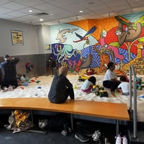 Children play in the Sandmagination indoor sandbox area with toys, while adults watch. A colorful mural with animals and children decorates the back wall. Shoes are lined up under a wooden bench in the foreground for the First Anniversary celebration.