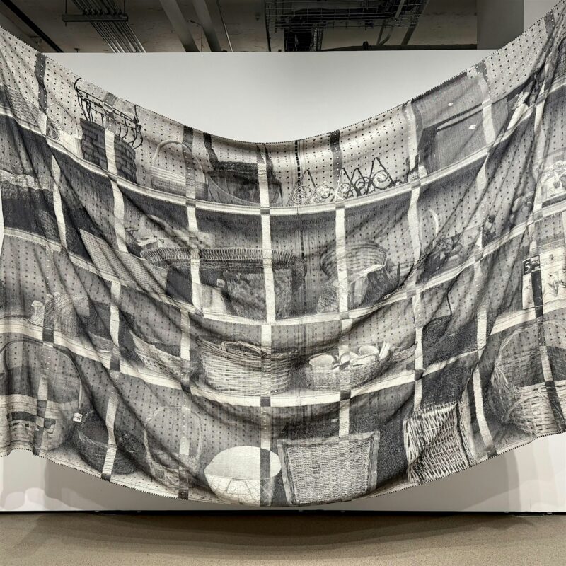 A large, semi-transparent fabric hangs in front of a white wall, printed with detailed black-and-white drawings of shelves filled with baskets, bowls, and various household items.
