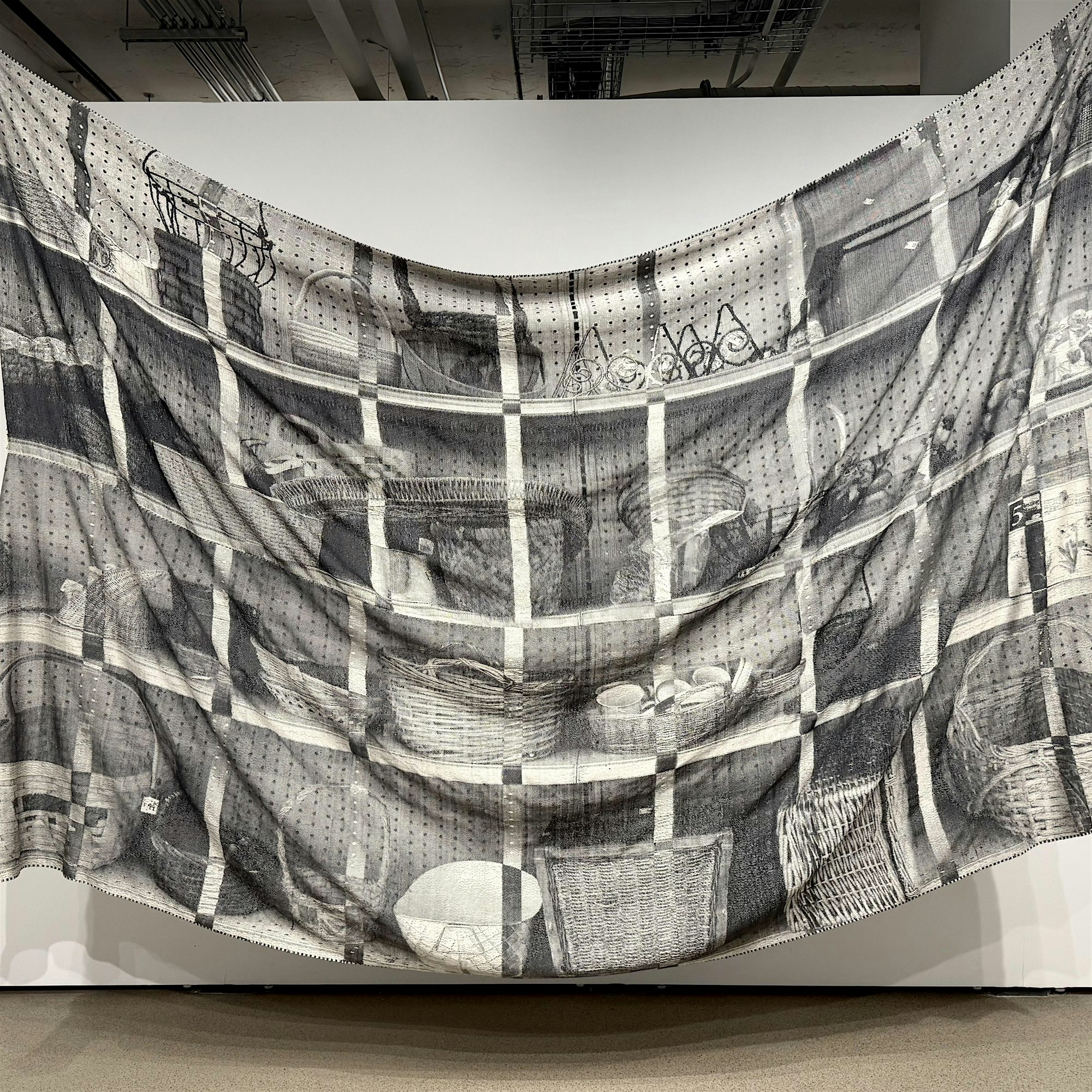 A large, semi-transparent fabric hangs in front of a white wall, printed with detailed black-and-white drawings of shelves filled with baskets, bowls, and various household items.