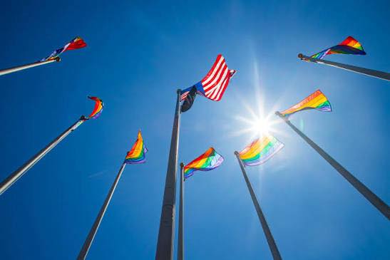 Multiple rainbow Pride flags and one American flag are displayed on flagpoles against a clear blue sky at the Salem Pride Flag Raising, with the sun shining brightly in the background—a vibrant highlight among Salem events.
