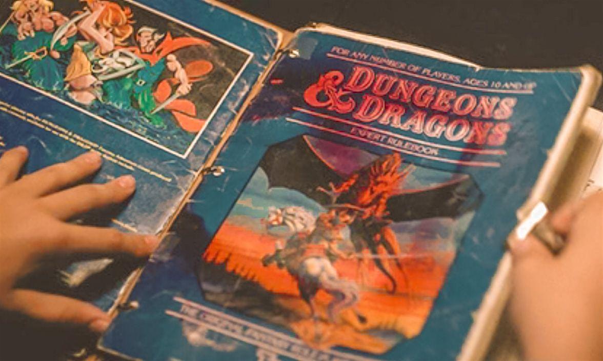 A person holds open a worn Dungeons & Dragons rulebook with a colorful illustrated cover featuring a dragon and a knight. The book is in a three-ring binder and shows signs of heavy use.
