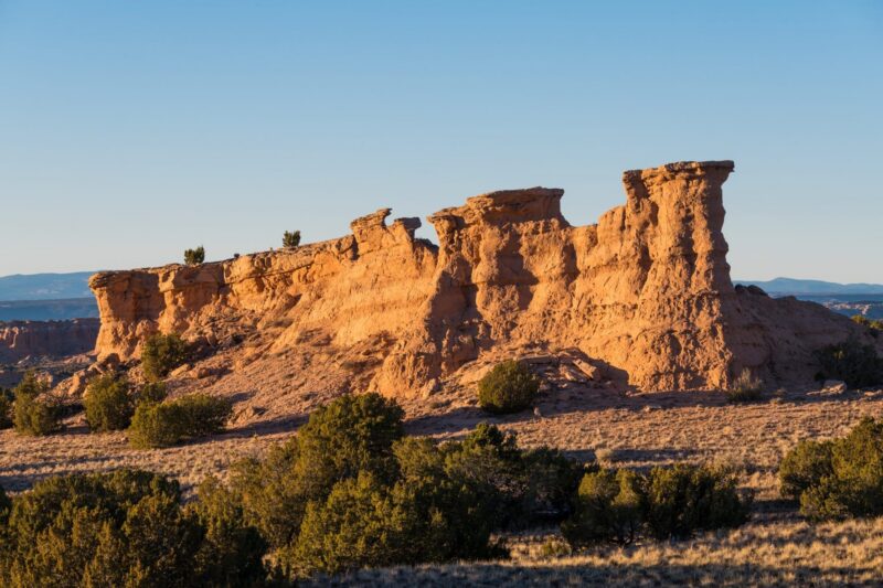 A rocky sandstone formation rises from the desert landscape, surrounded by sparse vegetation and shrubs under a clear blue sky in the late afternoon sunlight.