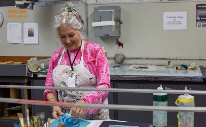 An older woman with gray hair wearing a pink tie-dye shirt and a paint-stained apron stands at a worktable, smiling as she works on an art project in a studio. Art supplies and tools are visible on the table.