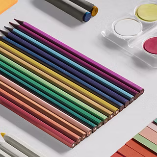 A neatly arranged set of colored pencils in various shades lies on a white surface, next to three round watercolor paints in a plastic case and other art supplies.
