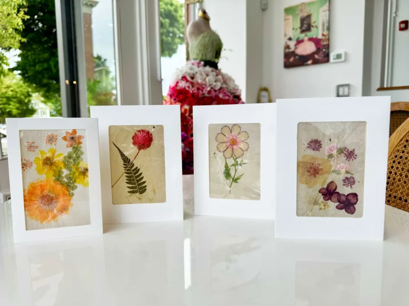 Four greeting cards featuring pressed flowers and leaves are displayed upright on a white surface in front of a window, echoing the creativity of a Botanical Workshop. A mannequin decorated with flowers and framed art are visible in the background.