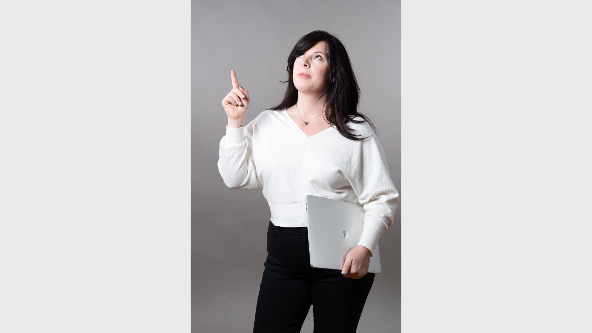Meet the Member: Andrea Ann Design—a woman with long dark hair, dressed in a white sweater and black pants, holds a closed laptop in her left hand and points upward with her right index finger against a plain gray background.