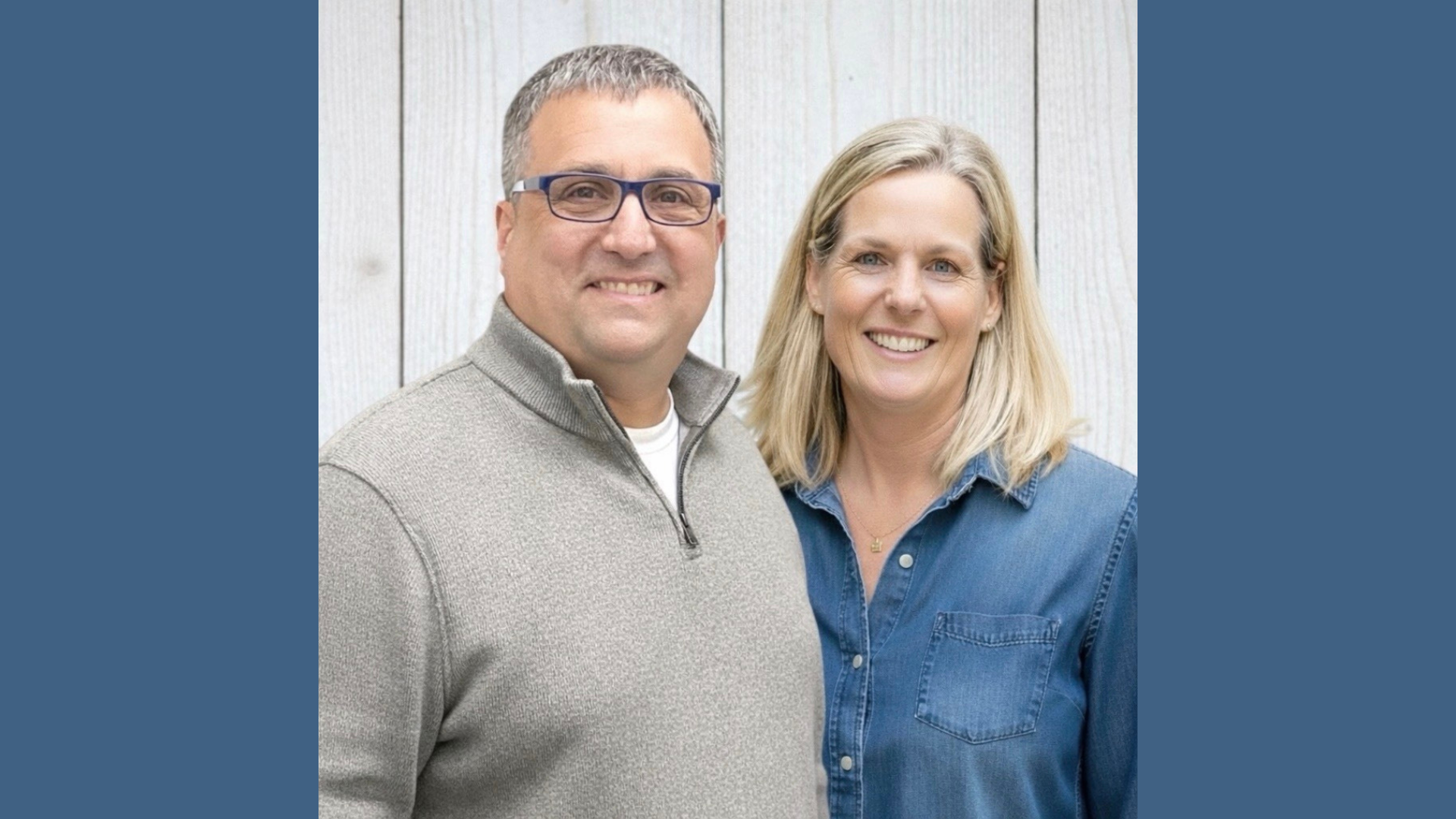 Meet the Member: Andrea Ann Design—a man and woman stand side by side, smiling at the camera. The man wears glasses and a gray zip-up sweater; the woman, with blond hair, sports a denim button-up shirt. A light wooden wall is in the background.