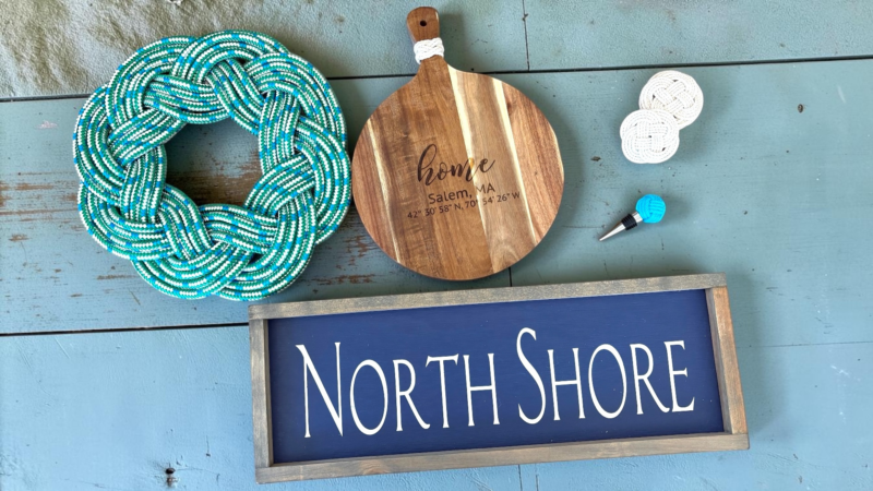 An arrangement of nautical-themed items by Odd Cod Studio on a blue wooden surface, featuring a turquoise rope knot, a wooden cutting board with text, a blue wine stopper, two round coasters, and a dark blue “North Shore” sign.