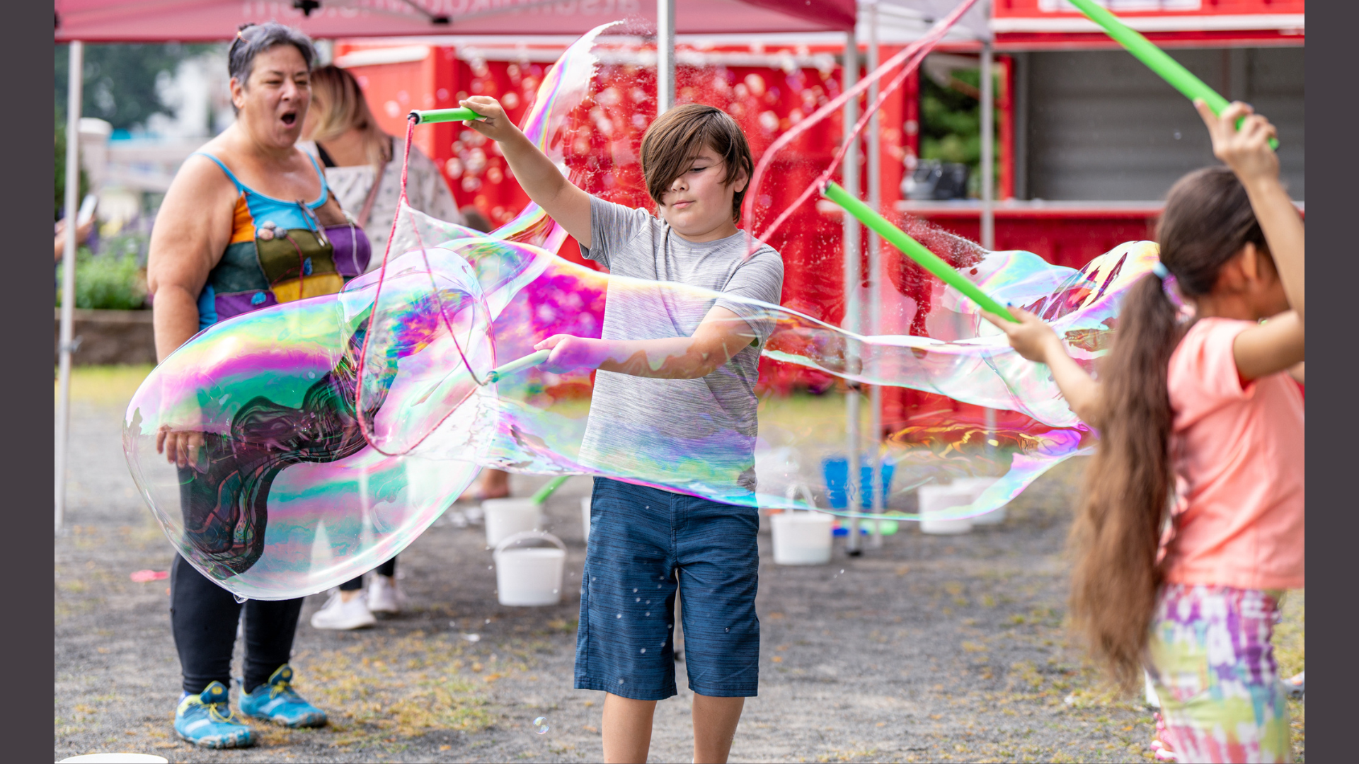A boy creates a large soap bubble using two sticks and a string, while a girl stands nearby holding similar sticks. In the background, an adult woman from Meet the Member: Airtight Garage watches under a red canopy.
