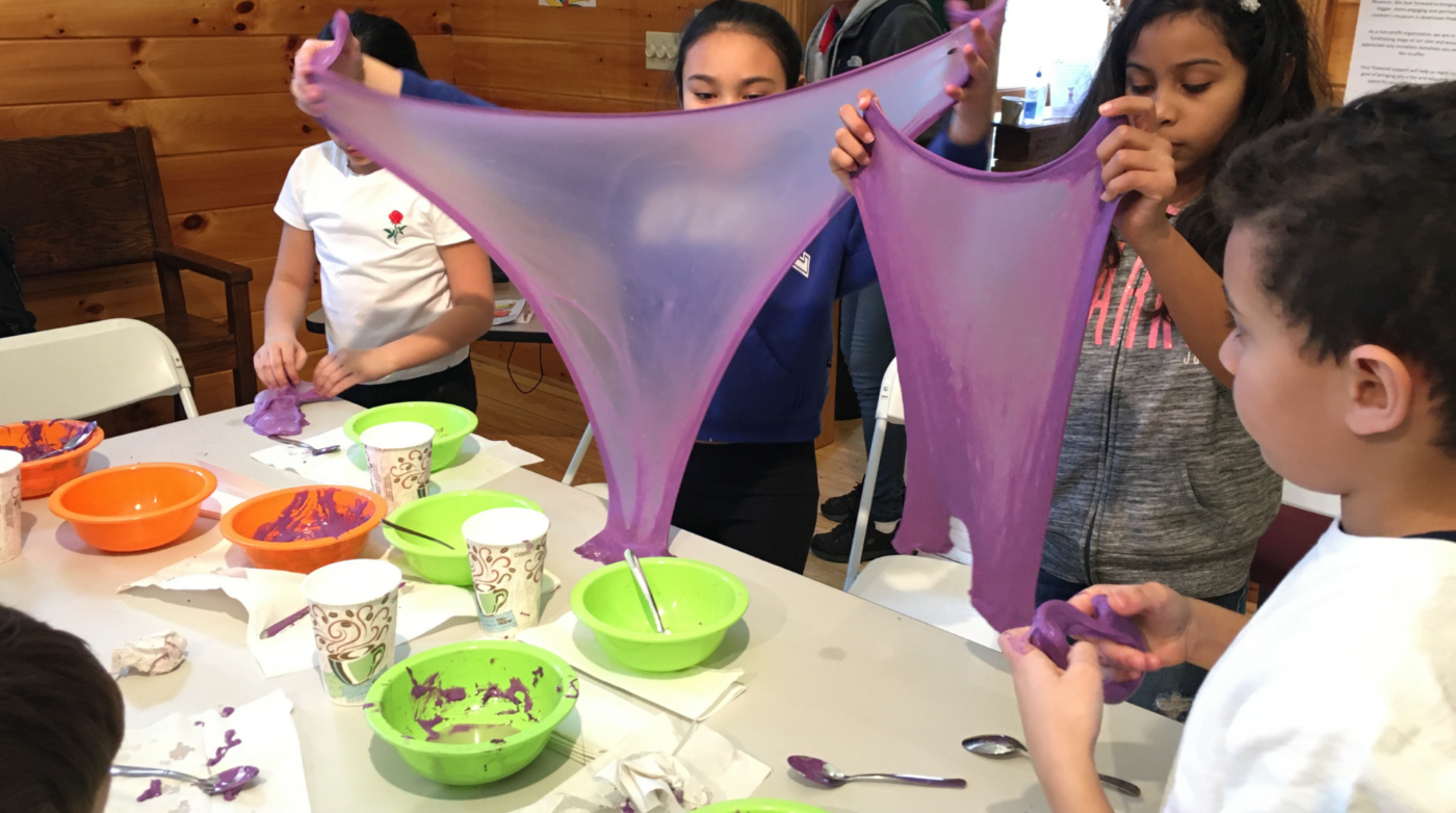 Children are stretching and playing with purple slime at a table covered with bowls, paper cups, and spoons in a room with wooden walls—an example of Outside the Box Learning that sparks creativity and hands-on education.