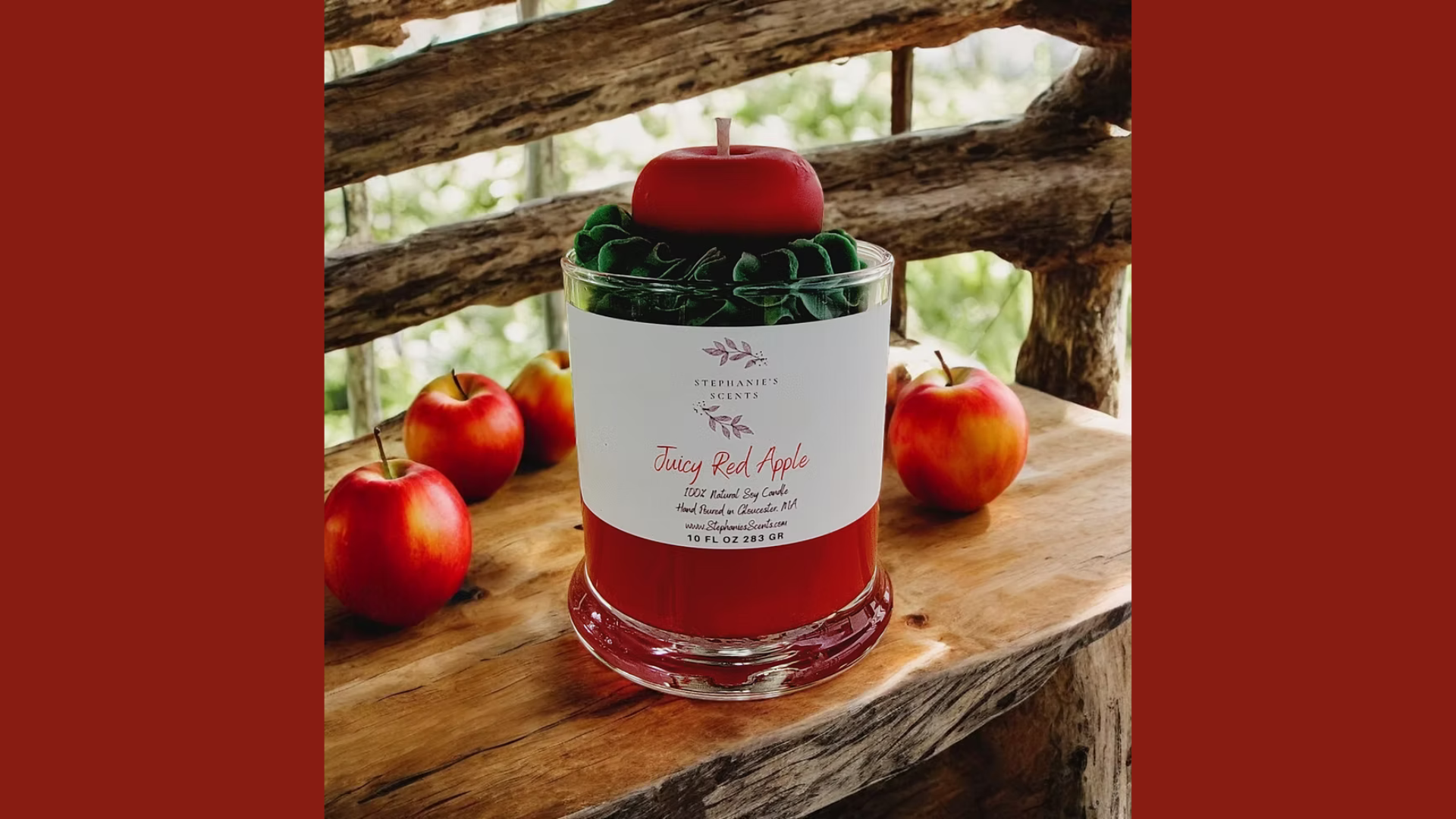 A red candle resembling an apple from Stephanie's Scents sits in a glass holder labeled "Juicy Red Apple," set on a wooden surface with four red apples and rustic branches, capturing the essence of premium fragrance.
