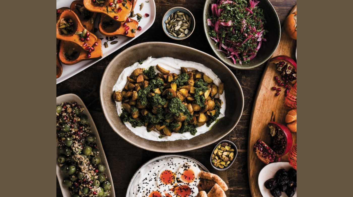 A variety of dishes are arranged on a table in true Flour in Her Hair style, including roasted squash quarters, salad, roasted vegetables on yogurt, stuffed grape leaves, sliced tomatoes with cheese, olives, nuts, and pomegranate seeds.