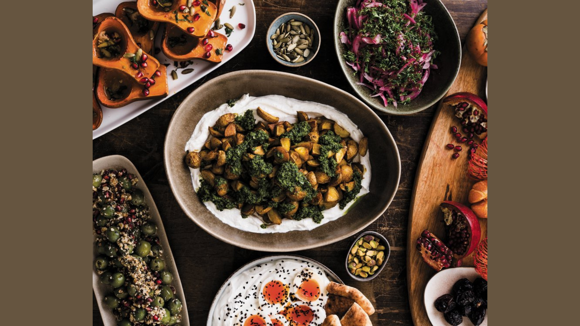 A variety of dishes are arranged on a table in true Flour in Her Hair style, including roasted squash quarters, salad, roasted vegetables on yogurt, stuffed grape leaves, sliced tomatoes with cheese, olives, nuts, and pomegranate seeds.