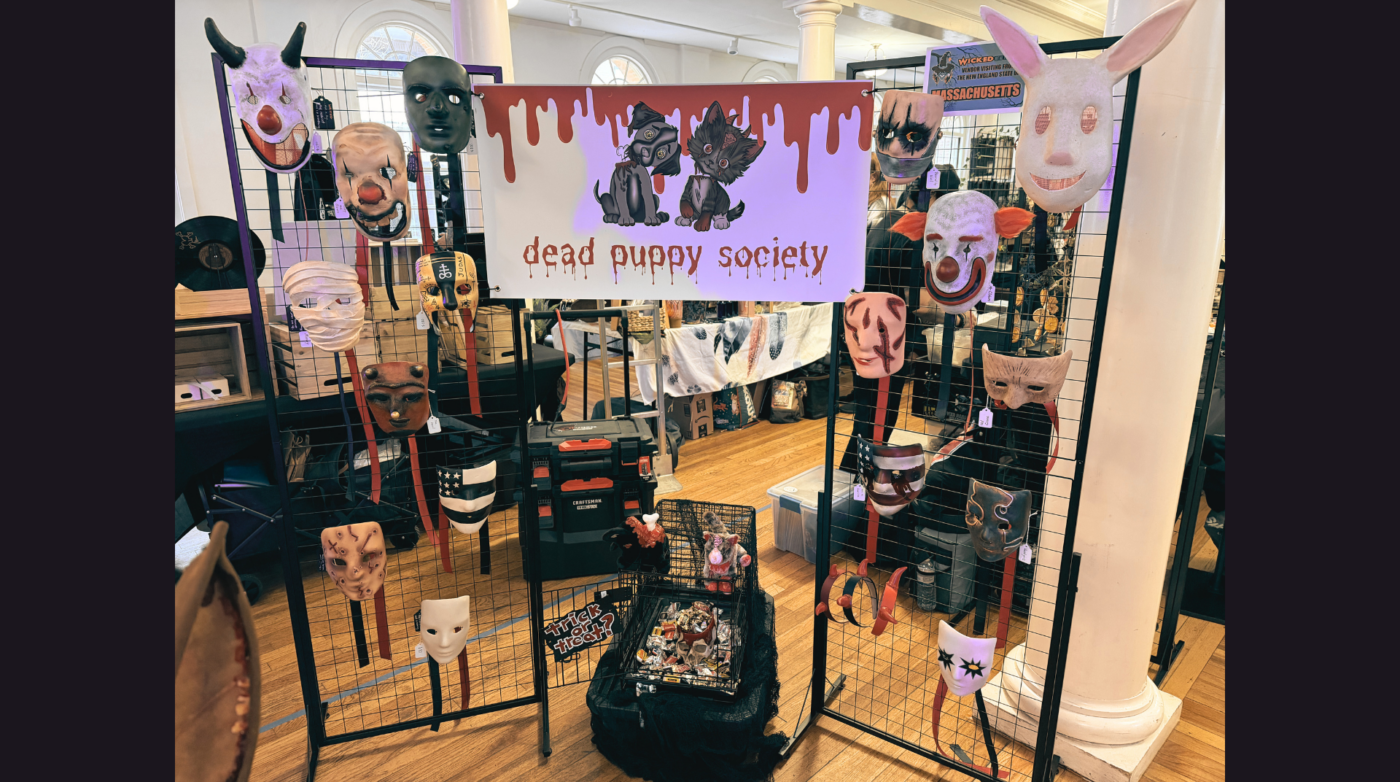 A display booth for Dead Puppy Society features animal and human masks on wire grids, a sign with illustrated dogs and dripping text, and a table with mask-making supplies in a well-lit indoor space.