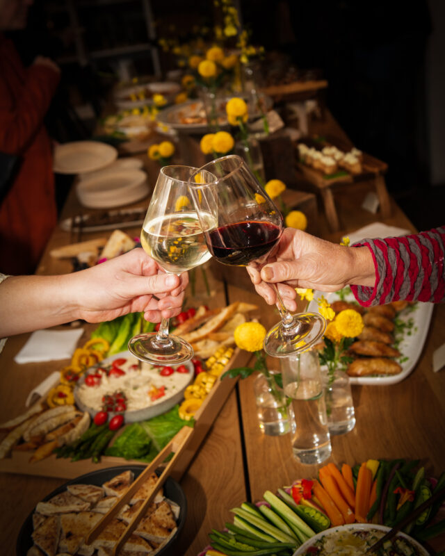 Two people clink wine glasses, one filled with white and the other with red, over a beautifully arranged dinner service featuring assorted appetizers, fresh vegetables, bread, and yellow flowers.