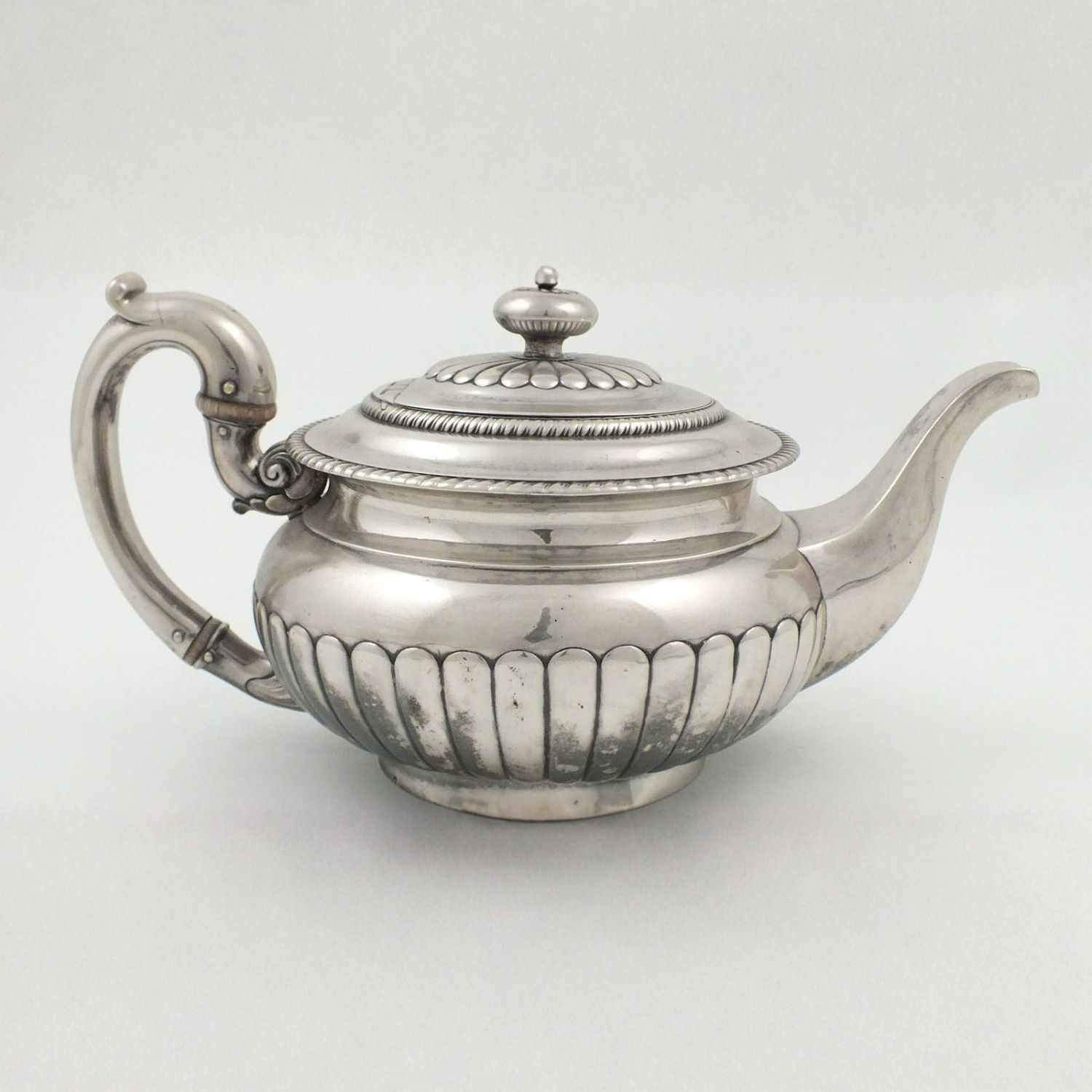 A silver teapot with a curved handle, a decorative lid with a knob, a ridged body, and a long spout, set against a plain light background.