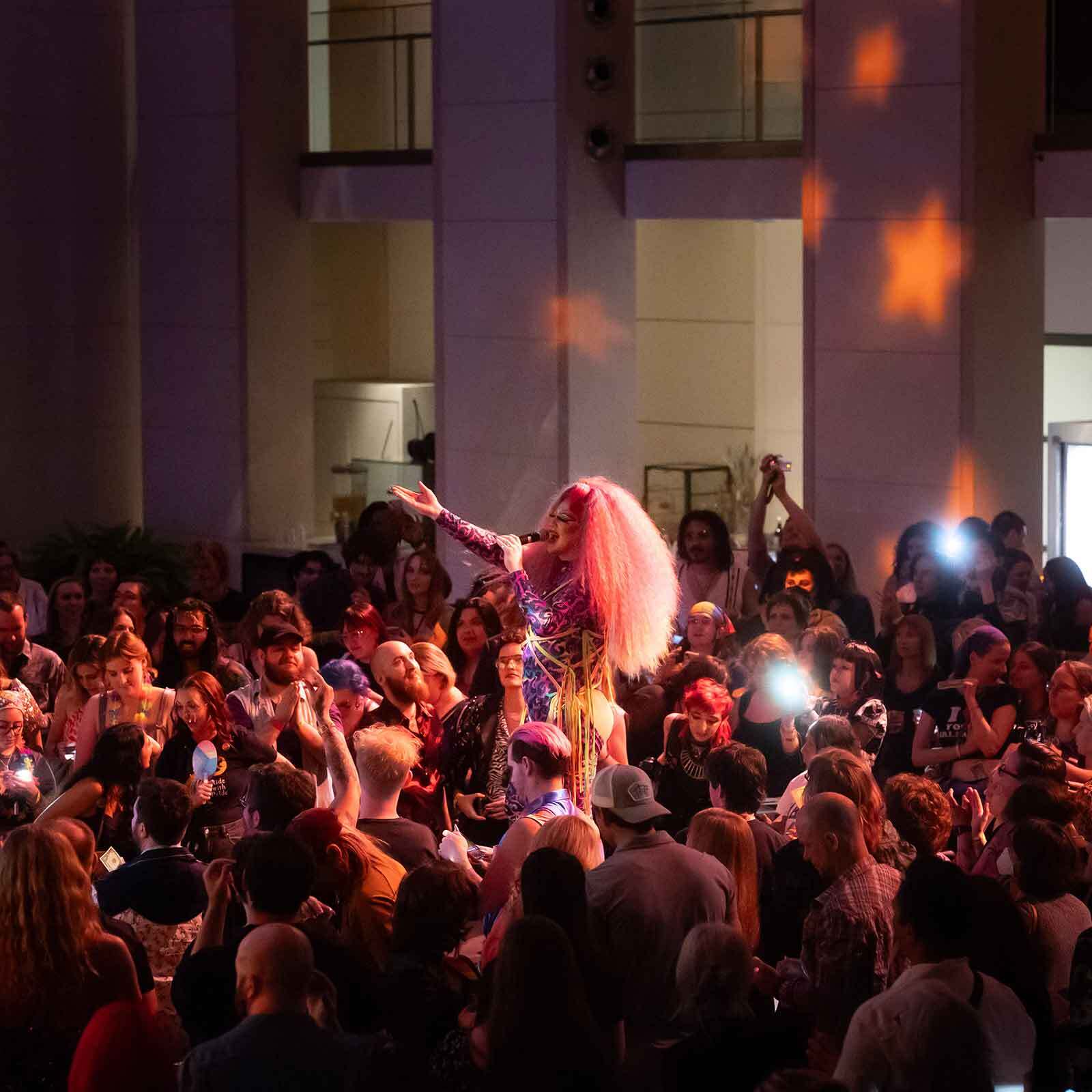 A performer in a colorful outfit with voluminous hair stands in the center of a crowd, reaching out with one arm. People surround the performer, some taking photos, in a large indoor space with light patterns on the walls.