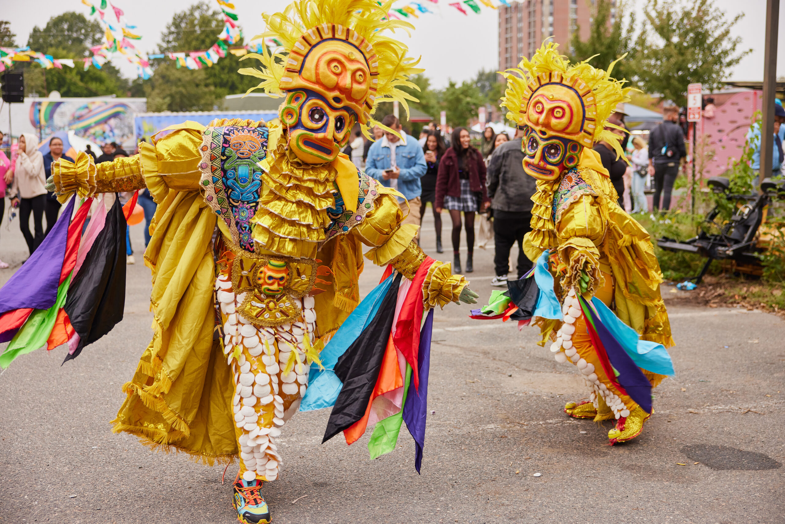 Two people wearing ornate yellow costumes and masks perform a traditional dance outdoors at a gathering. They hold colorful flags, entertaining a community of onlookers with trees and buildings visible in the background.